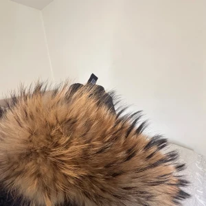 Svart pufferjacka Hollies med ”fuskpäls” - Snygg svart pufferjacka från Hollies med stor fluffig ”fuskpäls” på huvan. Jackan har dragkedja, knappar och coola detaljer som orange piping på insidan. Perfekt för kalla dagar och har en längre modell som håller dig varm hela vintern.