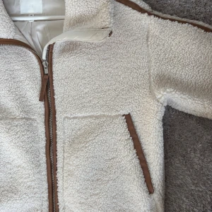 Vit teddyjacka från H&M med bruna detaljer - Säljer en supermjuk och fluffig vit teddyjacka från H&M med bruna detaljer längs dragkedja, fickor och ärmar. Perfekt för kyliga dagar när du vill vara både varm och snygg.☕️🍁🫶