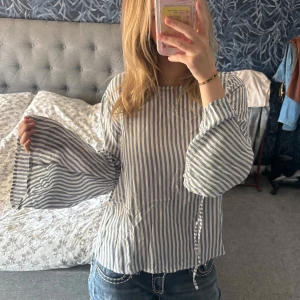 Randig blus med vida ärmar Pull&Bear - Säljer en snygg blus från Pull&Bear i storlek S. Den är vit med blåa vertikala ränder, har vida ärmar med knytband vid handlederna och en lös passform. Perfekt för dig som gillar en chill och trendig stil. Passar jättebra på sommaren, och sitter så skönt! 