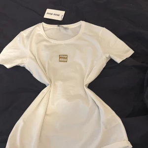 Vit Miu Miu t-shirt - Snygg vit t-shirt från Miu Miu med rund hals och korta ärmar. På bröstet sitter en stilren guldig Miu Miu-logga som ger en exklusiv vibe. T-shirten är gjord i mjuk bomull och har en klassisk passform som funkar till många olika looks. Fick som present men passar inte som jag önskar. (Vet inte om den r äkta eller inte men är rätt säker på att den är de)