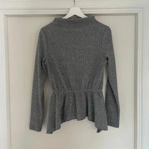 Grå ribbad peplumtröja från YAS - Supersnygg grå ribbad tröja från YAS med peplumvolang nedtill och lång ärm. Tröjan har en högre krage och markerad midja som ger en schysst siluett. Perfekt att styla med jeans eller kjol för en chill look.