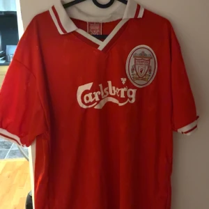 Liverpool retrotröjor Owen & Torres - Två klassiska Liverpool-matchtröjor med Carlsberg-tryck, en med Owen 10 och en med Torres 9 på ryggen. Båda är röda med vita detaljer, klubbmärke på bröstet och V-ringad krage. Perfekt för dig som älskar fotbollshistoria och vill ha något riktigt ikoniskt i garderoben.