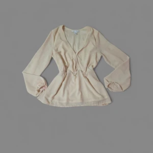 Beige omlottblus från NLY Trend - Supersnygg beige omlottblus från NLY Trend i storlek 38. Blusen har v-ringning, långa ärmar med resår vid handlederna och markerad midja. Tunt och lätt material som ger en flowy känsla. Perfekt att styla med jeans eller kjol för en trendig look.