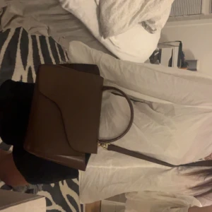 Brun handväska från ATP Atelier - Säljer en stilren brun handväska från ATP Atelier i slätt skinn. Väskan är i bra skick men skaftet är lite slitet annars funkar perfekt.  Ganska stor också så typ allt behövligt får plats. 