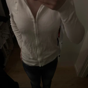 Vit kofta från Ralph Lauren med dragkedja - Säljer en klassisk vit kofta från Ralph Lauren med dragkedja framtill och diskret broderad logga på bröstet. Tröjan har långa ärmar, ribbade muddar och är gjord i mjuk bomull. Perfekt för lager-på-lager och enkel att matcha med jeans eller kjol.