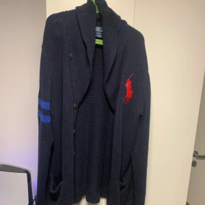 Mörkblå stickad kofta Polo Ralph Lauren - Snygg mörkblå stickad kofta från Polo Ralph Lauren med stora röda loggan på bröstet och blå ränder samt röd siffra 3 på ärmen. Koftan har knappar framtill samt 2 fickor. 