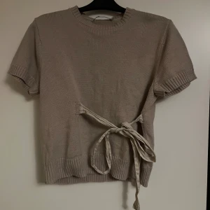 Beige stickad topp med knyt från Zara - Superfin beige stickad topp från Zara med korta ärmar och rund hals. Toppen har en snygg knyt-detalj i midjan som ger en extra touch. Perfekt till jeans eller kjol för en chill och trendig look.