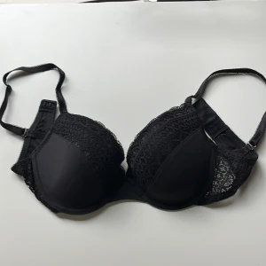 Svart spets-bh från Twilfit 70D - Snygg svart bh från Twilfit i storlek 70D med fina spetsdetaljer och justerbara axelband. Kuporna är vadderade och har en elegant spets längs kanterna och sidorna. Perfekt för dig som gillar en klassisk look med lite extra detaljer.