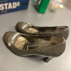 Grå vintage quiltade klackarskor med dragkedjedetalj - Snygga vintage coola grå klackarskor från Joy & Peace med quiltat mönster och silverfärgad dragkedjedetalj med hjärtberlock framtill. Skorna har rund tå och låg klack, tillverkade i glansigt syntetmaterial. Perfekta för dig som gillar unika detaljer och klassisk stil.