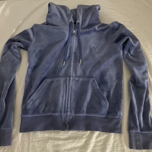 Blå velour hoodie från Juicy Couture - Säljer en blå velour hoodie från Juicy Couture med dragkedja och huva. Tröjan har långa ärmar, känguruficka framtill och broderad logga på bröstet. Materialet är mjuk polyesterblandning som ger en lyxig känsla. Perfekt för en chill och trendig look. Köpt för 600kr