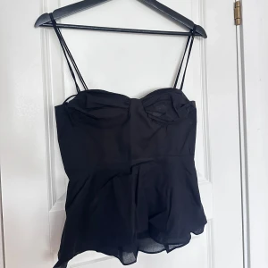 Svart topp med tunna axelband - Den populära Zara toppen med tunna dubbla axelband och lätt volang nertill. Toppen har markerade kupor och smockad rygg för extra komfort. Perfekt att styla med jeans eller kjol för en trendig look.