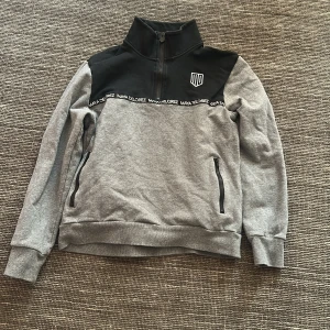 Grå half zip-tröja Maya Delorez M - Snygg grå tröja från Maya Delorez med svart axelparti och broderad logga. Half zip-dragkedja framtill, två fickor med dragkedja och ribbade muddar. Perfekt för dig som gillar sportig och clean stil.