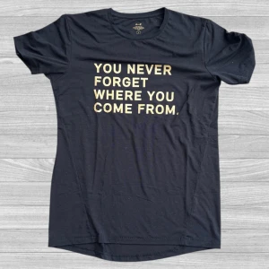 Svart t-shirt med tryck från A–Z - Svart t-shirt från A–Z i storlek L med stort gult texttryck på bröstet: 'YOU NEVER FORGET WHERE YOU COME FROM.' Klassisk rund hals och korta ärmar. Skön och mjuk bomullskvalitet, perfekt till jeans eller shorts.
