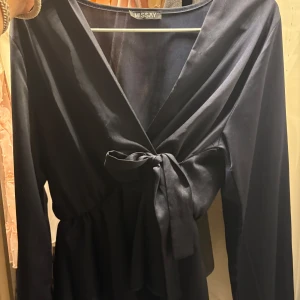 Marinblå omlottklänning från Missay - Snygg marinblå omlottblus från Missay med lång ärm och knyt i midjan. Blusen har en v-ringad design och är tillverkad i ett mjukt, glansigt material som ger en elegant look. Perfekt för dig som gillar stilrena och klassiska plagg.