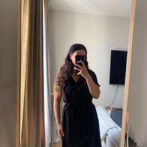 Svart kort klänning från H&M - Säljer en svart kort klänning från H&M med v-ringning och knyt i midjan. Klänningen har korta, lite fladdriga ärmar och är tillverkad i ett lätt och mjukt syntetmaterial. Perfekt för dig som gillar enkel och stilren look.