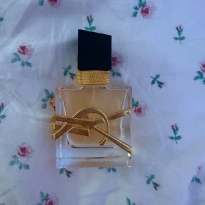 Yves Saint Laurent Libre Eau de Parfum - Säljer en lyxig parfym från Yves Saint Laurent, Libre Eau de Parfum. Flaskan är fyrkantig i genomskinligt glas med en elegant guldig YSL-logga och svart kantig kork med gulddetalj. Doften är modern och ikonisk, perfekt för dig som gillar exklusiva parfymer.