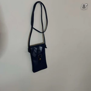 Blå axelväska i skinn - Snygg blå axelväska i skinn med lång rem och lock med spänne. Väskan har en stilren och minimalistisk design, perfekt för att bära mobilen och småsaker. Locket har en lätt glansig finish som ger en cool touch.