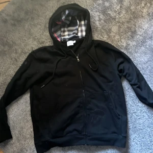 Svart Burberry hoodie med rutigt foder - Svart hoodie från Burberry i storlek M med dragkedja och klassiskt rutigt foder i huvan. Tröjan har snörning vid huvan, två fickor framtill och är långärmad. Perfekt för dig som gillar stilrena och exklusiva streetwear-plagg.