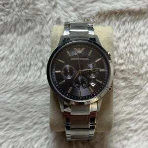 Emporio Armani silverfärgad klocka - Stilren klocka från Emporio Armani med svart urtavla och silverfärgat armband i metall. Klockan har kronograf-funktion, datumvisning och klassisk rund form. Levereras med originalask och extra länk. Perfekt för dig som gillar snygga accessoarer.