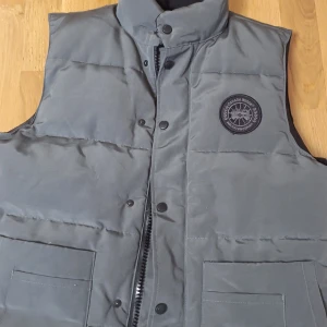Grå dunväst från Canada Goose - Säljer en grå dunväst från Canada Goose med hög krage, två stora fickor framtill och diskret logga på bröstet. Västen har både dragkedja och knappar framtill och är fodrad med mjukt material. Perfekt för kyliga dagar när du vill hålla stilen.