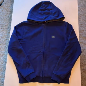 Blå hoodie från Lacoste Sport - Säljer en blå hoodie från Lacoste Sport med dragkedja och huva. Klassisk krokodil-logga på bröstet och ribbade muddar vid ärmslut och nederkant. Perfekt för dig som gillar sportig och stilren look.