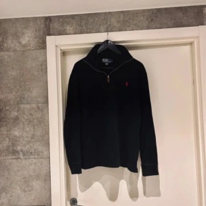 Svart stickad half zip från Polo Ralph Lauren - Svart stickad tröja från Polo Ralph Lauren med röd broderad logga på bröstet. Tröjan har half zip med läderdetalj på dragkedjan och hög krage. Perfekt för dig som gillar klassisk och stilren look. Storleken är S, sitter perfekt om du är 185-175cm.