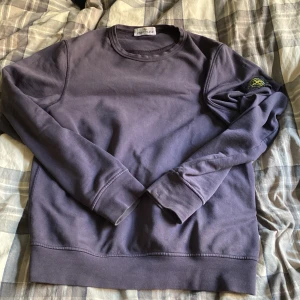 Mörklila sweatshirt från Stone Island - Snygg mörklila sweatshirt från Stone Island Junior med klassisk rund hals och den ikoniska patchen på vänster ärm. Tröjan är tillverkad i mjuk bomull och har långa ärmar med ribbade muddar. Perfekt för dig som gillar streetwear och vill ha en clean look.