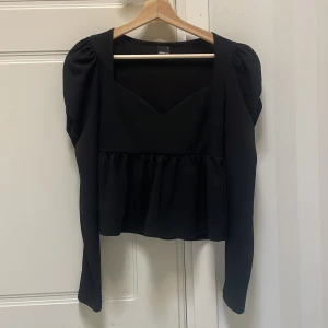 Svart blus med puffärm från Gina Tricot - Supersnygg svart blus från Gina Tricot med puffiga axlar och lång ärm. Toppen har en markerad midja med rynk och en v-ringad urringning framtill. Perfekt för dig som gillar trendiga detaljer och stilren look. Använd en gång under ett nyårsfirande, säljer då jag upplever att den satt lite tajt. 