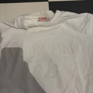 Vit Valentino t-shirt med relieftryck - Vit t-shirt från Valentino med stilrent relieftryck på bröstet där det står 'VALENTINO Maison Established 1960' och logotypen. Klassisk rund hals och korta ärmar. Mjuk och skön bomullskvalitet, perfekt för en clean och snygg look.