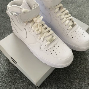 Nike Air Force 1 - Nike Air Force 1 Mid sneakers i helvitt skinn med klassisk snörning, perforerad tå och kardborreband runt ankeln. Snygg och tidlös modell med rund tå och platt sula, perfekt för dig som gillar en clean och sportig look.