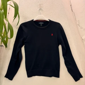 Svart tröja från Polo Ralph Lauren - Klassisk svart tröja från Polo Ralph Lauren i mjuk pima-bomull. Tröjan har rund halsringning, långa ärmar och ikonisk röd broderad logga på bröstet. Perfekt för en clean och stilren look.