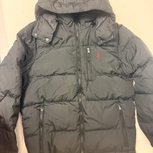 Svart pufferjacka från Polo Ralph Lauren storlek S_M - Svart pufferjacka från Polo Ralph Lauren med huva, dragkedjor på fickor och bröst, samt röd logga på vänster sida. Jackan är tillverkad i återvunnen polyester och har en vattenavvisande yta. Perfekt för kalla dagar med sin tjocka och quiltade design.
