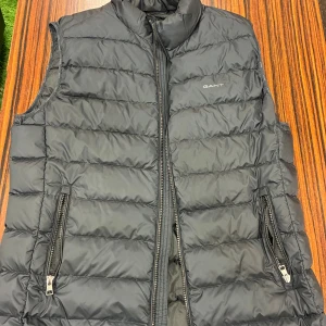 Svart dunväst från Gant - Svart quiltad dunväst från Gant med hög krage och två dragkedjeförsedda fickor. Västen har en stilren design med diskret logga på bröstet och hel dragkedja framtill. Perfekt för lager-på-lager under kyliga dagar.