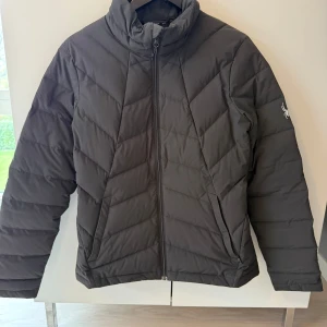 Svart pufferjacka från Spyder - Snygg svart pufferjacka från Spyder med coolt spindeltryck på ärmen och logga på ryggen. Jackan har quiltad design, hög krage och dragkedja framtill. Perfekt för kalla dagar och har en modern, sportig look.