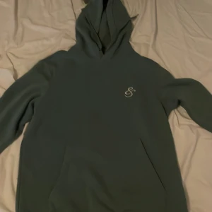 Grön hoodie med broderad logga - Mysig grön hoodie från SikSilk med broderad logga på bröstet. Tröjan har huva, långa ärmar och en klassisk känguruficka framtill. Perfekt för chill dagar eller när du vill ha en enkel streetstyle-look.