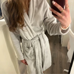 Ljusgrå fleece morgonrock med bälte - Supermjuk ljusgrå morgonrock i fleece med långa ärmar, stora fickor och knytskärp i midjan. Perfekt för att chilla hemma och hålla sig varm. Klassisk omlottmodell med skön känsla mot huden.