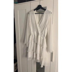 Vit omlottklänning med volanger - Superfin vit kortklänning med omlottliv, långa ärmar och flera lager volanger i kjolen. Klänningen har v-ringning och ett lätt, luftigt tyg som ger en romantisk känsla. Perfekt för sommarens alla tillfällen!