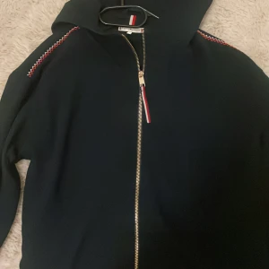 Mörkblå hoodie från Tommy Hilfiger - Mörkblå  hoodie från Tommy Hilfiger med dragkedja i guld och snygga röda och vita detaljer på axlarna. Klassisk huva och långärmad modell, med diskret logga på ärmen och knappar vid ärmslut. Perfekt för en chill och stilren look. Den är i storlek 8 o passar S/M