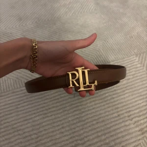 Brunt skärp Ralph Lauren Inspo - Snyggt brunt bälte Ralph Lauren logga med ett stort guldfärgat RLL-spänne. Bältet är gjort i slätt skinn och har en klassisk, stilren design som passar till många outfits.