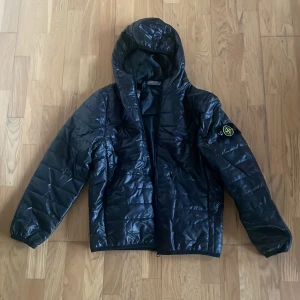 Svart pufferjacka från Stone Island - Svart pufferjacka från Stone Island med huva och klassisk patch på ärmen. Jackan har quiltad design, dragkedja framtill och är tillverkad i glansig syntet. Perfekt för dig som vill ha en clean och stilren look med street-vibe.