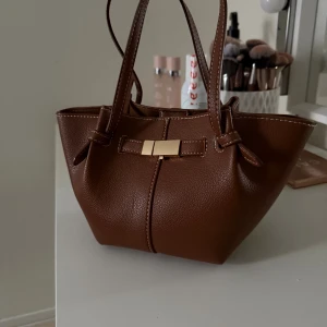 Brun handväska med gulddetalj - Snygg brun handväska i skinnimitation med två handtag och en elegant guldfärgad detalj framtill. Väskan har en stilren form och är perfekt för att bära dina viktigaste saker. Insidan är rymlig och stängs med magnetknapp. Passar till många olika outfits.