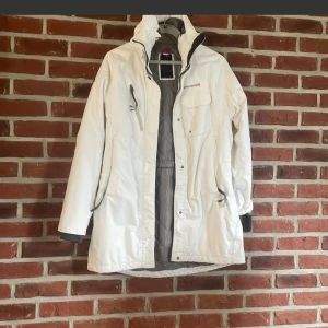 Vit parkasjacka från Didriksons 1913 - Snygg vit parkasjacka från Didriksons 1913 med grått quiltat foder och Storm System-detaljer. Jackan har huva, flera fickor med dragkedja och tryckknappar, samt justerbara muddar. Perfekt för kalla dagar och har en clean, sportig look.