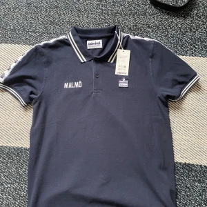 Mörkblå pikétröja Admiral Malmö M. Aldrig använd endast testa den. Nypris: 600 kr mitt pris 350 kr - Snygg mörkblå pikétröja från Admiral med vita ränder på krage och ärmslut. Broderad text 'Malmö' på bröstet och Admiral-logga. Klassisk piké med knappslå och sportig känsla, perfekt för dig som gillar stilren streetwear.