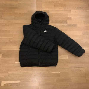 Svart pufferjacka från Nike XL - Svart pufferjacka från Nike i storlek XL med huva och dragkedja. Jackan har ett quiltat mönster och Nike-logga på bröstet. Insidan har en orange namnlapp. Perfekt för kalla dagar och har en loose passform.