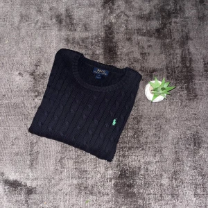 Svart kabelstickad tröja Polo Ralph Lauren - Kabelstickad svart tröja från Polo Ralph Lauren med rund halsringning och klassisk grön broderad logga på bröstet. Tröjan är i bomull och har långa ärmar samt ribbade muddar för en snygg passform.