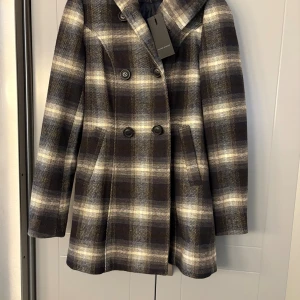 Rutig kappa med huva från Vero Moda - Snygg rutig kappa från Vero Moda i storlek S. Jackan har dubbelknäppning med svarta knappar, huva och två fickor fram. Färgerna går i mörkblått, grått och beige. Materialet är ullblandning som håller dig varm under höst och vinter.