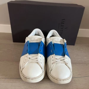 Valentino Garavani Open sneakers blå/vit - Snygga Valentino Garavani Open sneakers i vitt skinn med en bred blå läderdetalj över mitten. Klassisk rund tå, vita snören och ikoniska nitar baktill på sulan. Kommer med originalkartong, dustbags och extra skosnören.