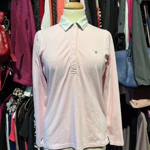 Ljusrosa pikétröja från Gant - Snygg ljusrosa pikétröja från Gant med vit krage och diskret logga på bröstet. Tröjan har långa ärmar, klassisk pikéknäppning och små slitsar i sidorna.