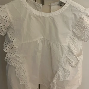 Vit blus med volang och broderi - Superfin vit blus med korta ärmar och broderade volangdetaljer längs axlar och sidor. Blusen har rund halsringning och är tillverkad i bomull, vilket gör den luftig och perfekt för varmare dagar. Söt och trendig med öppen detalj i nacken.