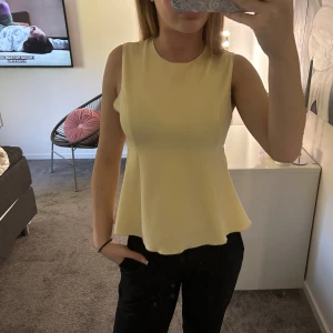 Gul ärmlös topp med utsvängd midja - Snygg ljusgul ärmlös topp med rund halsringning och utsvängd midja som ger en feminin siluett. Toppen är enkel och stilren, perfekt att matcha med både jeans och kjol. Materialet känns mjukt och följsamt mot kroppen.